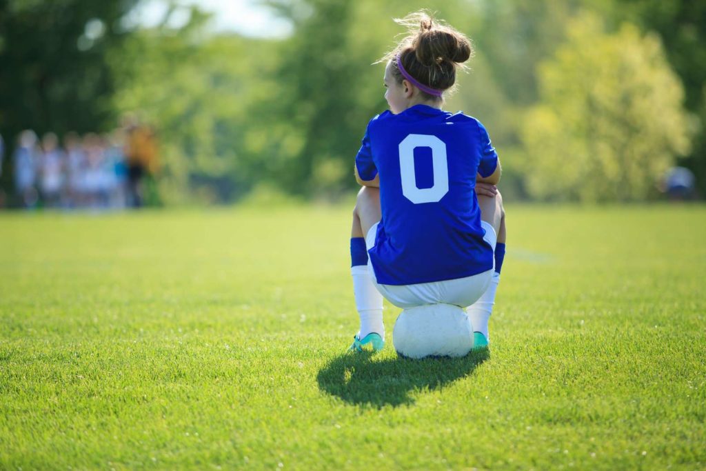 En flicka sitter på en boll på en fotbollsplan. Fysisk aktivitet och ansträngning kan utlösa ett astmaanfall – kanske var hon tvungen att ta en paus.