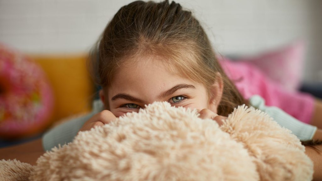 En flicka kikar upp över sin teddybjörn. Vid kvalsterallergi bör mjuka leksaker tvättas regelbundet i tvättmaskinen för att minska allergenexponeringen.