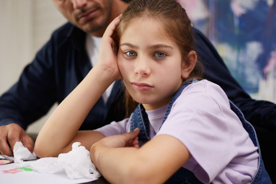 En flicka sitter vid ett bord och ritar tillsammans med sin pappa. Hon ser trött ut och stöder huvudet i handen. Hon kanske lider av trötthet och koncentrationssvårigheter på grund av pollenallergi.