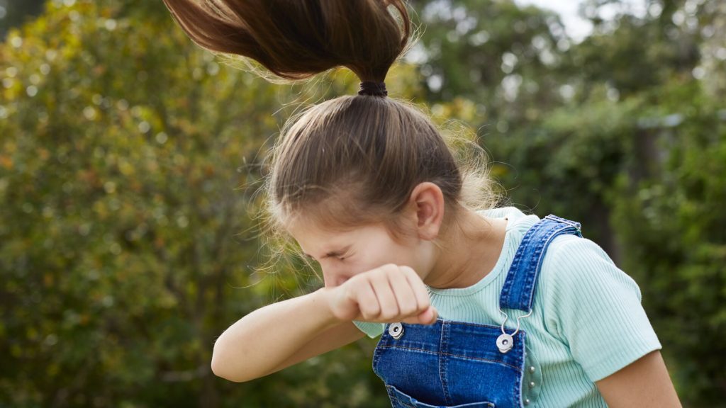 En flicka med hästsvans och klädd i blå klänning nyser i sin armbåge. Hon är omgiven av buskar och träd som sprider pollen och utlöser en allergisk reaktion.